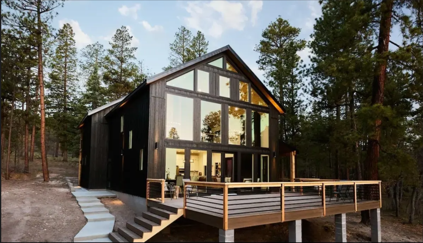 Modern black home with expansive windows and a wood deck surrounded by pine trees